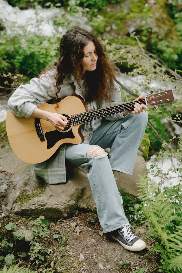 about-us Young woman strumming an acoustic guitar outdoors, surrounded by greenery.