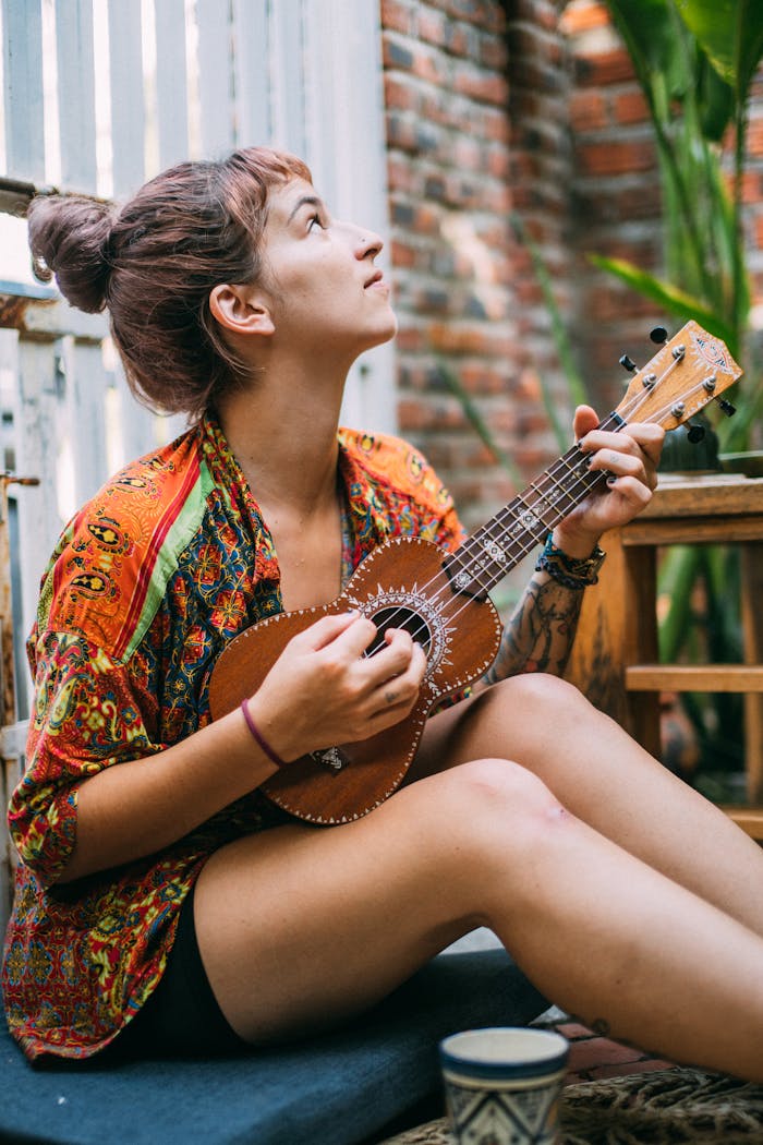 A woman sitting and playing a ukulele in an indoor setting, looking up thoughtfully.